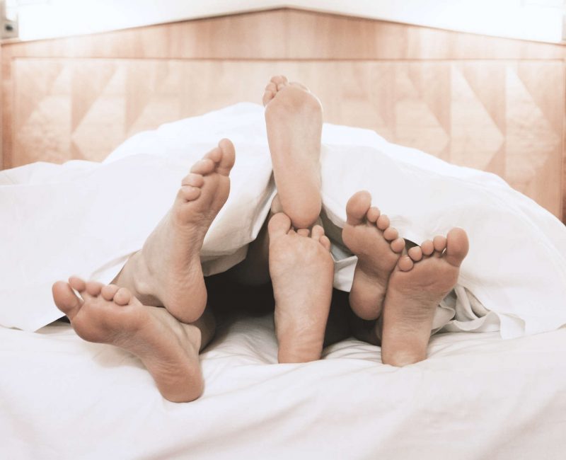 Three,Pairs,Of,Feet,Lying,Together,Under,Bed,Cover,In